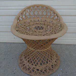 Puede incluir: Silla de mimbre marrón claro con asiento redondo y respaldo curvo. La silla tiene un diseño tejido y una base acampanada. La silla está hecha de materiales naturales y tiene un aspecto vintage.