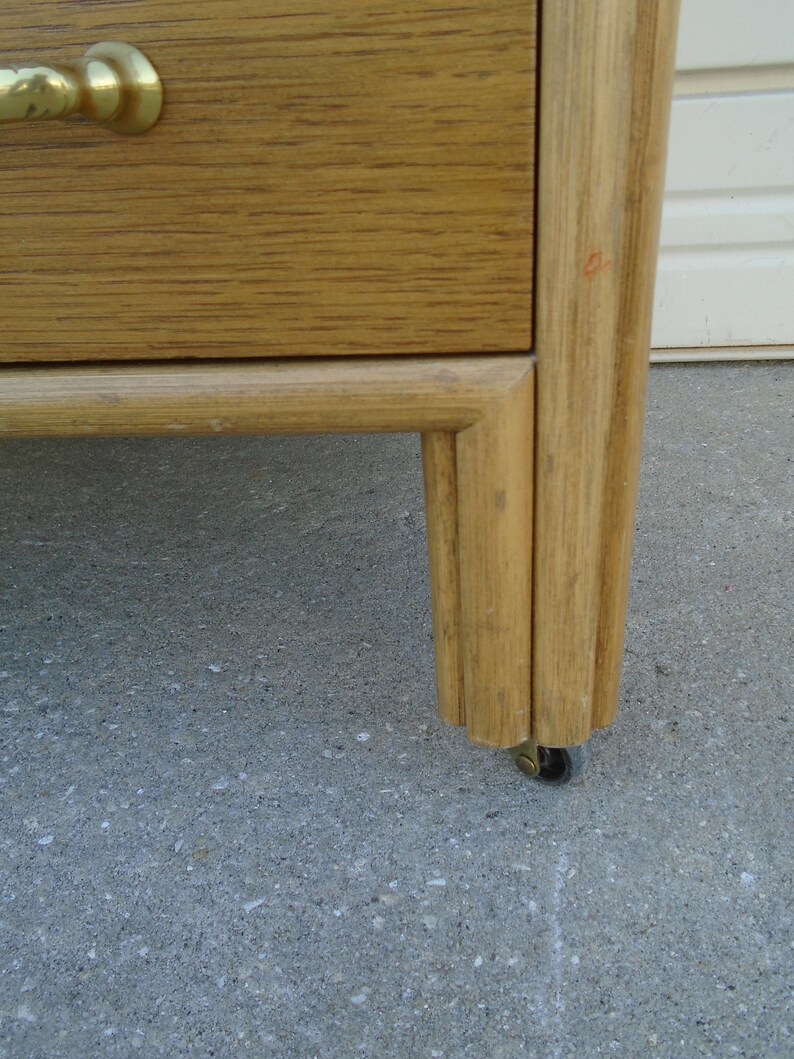 Puede incluir: Primer plano de una c&oacute;moda de madera marr&oacute;n claro con un tirador de lat&oacute;n. La c&oacute;moda tiene un caj&oacute;n y est&aacute; sostenida por una pata con una peque&ntilde;a rueda. La veta de la madera es visible.