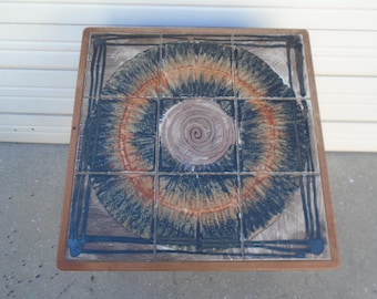 May include: A square wooden table top with a ceramic tile inlay. The tile is a mosaic of blue, orange, and brown colors with a spiral design in the center.