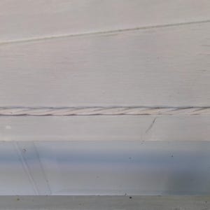May include: Close-up of a section of a white painted wooden structure. A twisted rope-like detail runs horizontally across the frame, adding a textural element to the design. The wood grain is visible.