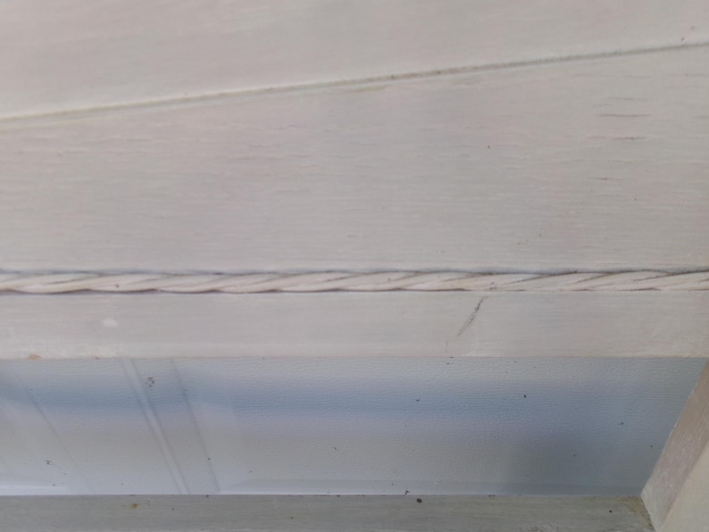 May include: Close-up of a section of a white painted wooden structure. A twisted rope-like detail runs horizontally across the frame, adding a textural element to the design. The wood grain is visible.
