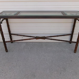May include: A dark brown console table with a glass top and bamboo-style legs. The table has a rectangular shape with a glass insert in the center. The legs are angled and connected by crossbars for stability. The table is approximately 48 inches long.
