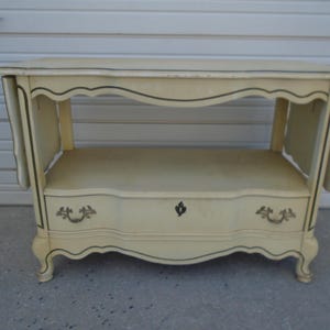May include: A white painted wooden server with a single drawer and a shelf. The server has a curved, ornate design and gold accents.