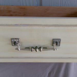 May include: Close-up of a drawer front with a pale yellow finish and a silver handle. The handle has a decorative design with square backplates and a central bar. The drawer's interior is visible, showing natural wood.