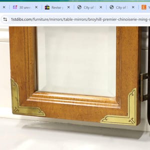 May include: Close-up of a wooden framed mirror with gold accents. The mirror has a beveled edge and is attached to a larger piece of furniture.