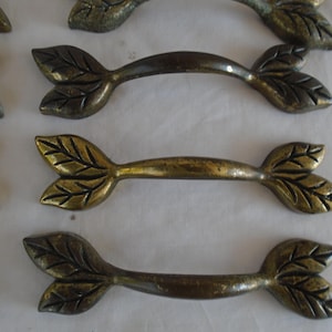 May include: Set of eight antique brass cabinet pulls with a leaf design. Each pull has a curved handle and two leaf-shaped ends.