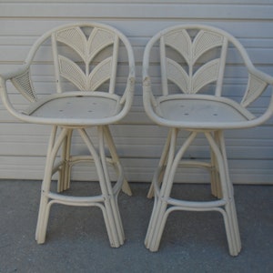 May include: Two white wicker bar stools with a leaf design on the back. The stools have a white padded seat and a white frame.