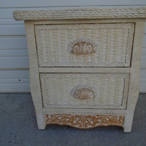 May include: A two-drawer wicker nightstand in a cream color. The drawers have decorative handles and are woven. The bottom of the nightstand has a decorative wood trim. The nightstand has a woven top.