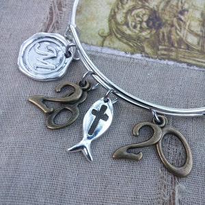 May include: Silver bangle bracelet with multiple charms. Charms include a silver initial seal, a fish with a cross, and two bronze-colored number 20 charms. The bracelet is displayed on a textured, neutral-colored surface.
