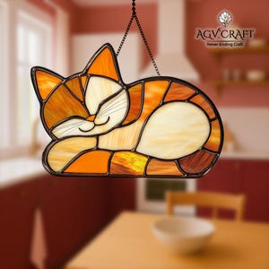 May include: Stained glass cat suncatcher in shades of orange, cream, and brown. The cat is in a curled-up sleeping position, with a smiling face and black whiskers. The suncatcher is hanging from a chain. The text "AGV Craft Never Ending Craft" is visible.