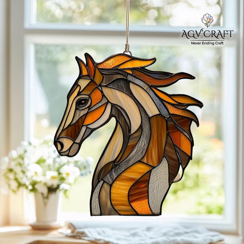 Peut inclure: Suncatcher en forme de t&ecirc;te de cheval en vitrail aux tons marron, orange et beige. Le cheval est suspendu par un fin cordon beige. Le logo AGV Craft est visible en haut &agrave; droite. Le capteur solaire est pr&eacute;sent&eacute; devant une fen&ecirc;tre.