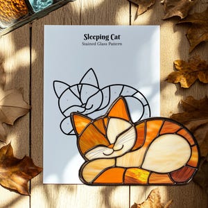 May include: A stained glass cat in shades of orange, yellow, and cream, resting on a white paper with a pattern outline. The paper reads "Sleeping Cat Stained Glass Pattern". The cat is surrounded by autumn leaves on a wooden surface.