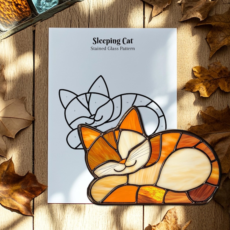 May include: A stained glass cat in shades of orange, yellow, and cream, resting on a white paper with a pattern outline. The paper reads "Sleeping Cat Stained Glass Pattern". The cat is surrounded by autumn leaves on a wooden surface.