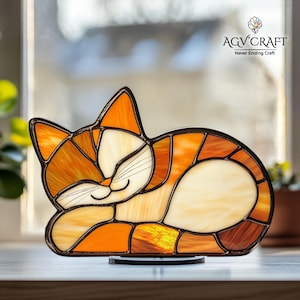 May include: A stained glass cat figurine in shades of orange, cream, and brown, with a black outline. The cat is depicted sleeping, with its eyes closed and a gentle smile. The figurine is on a black base, with the text "AGV CRAFT Never Ending Craft".