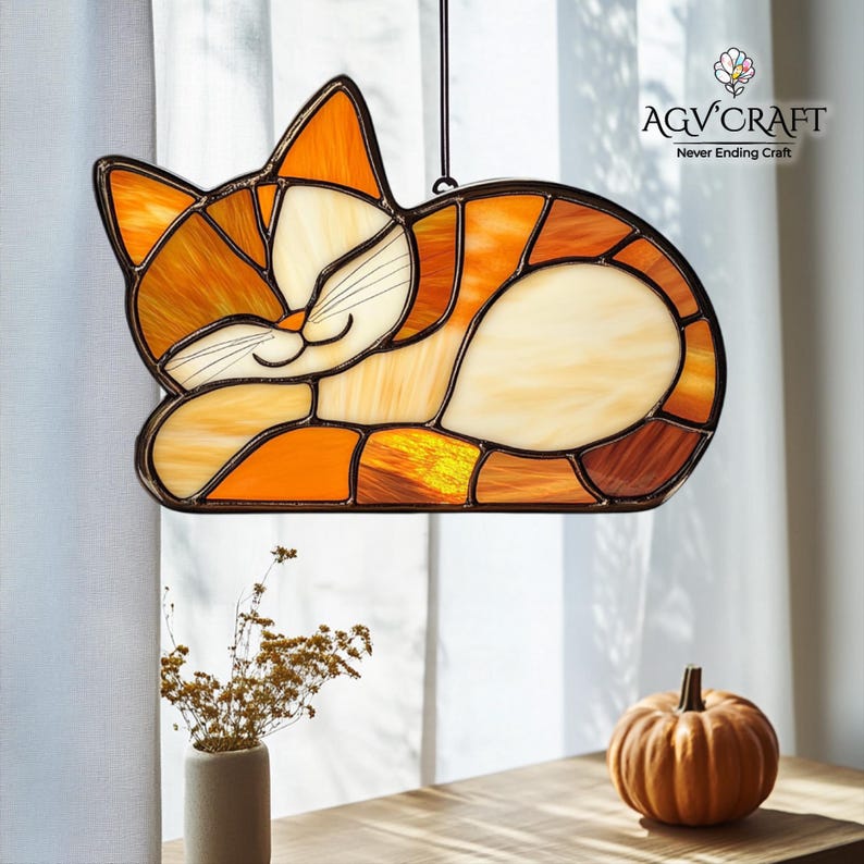 May include: Stained glass cat suncatcher in shades of orange, cream, and brown. The cat is depicted in a sleeping pose with a smiling face. The AGV Craft logo is visible in the upper right corner. A decorative item for cat lovers.