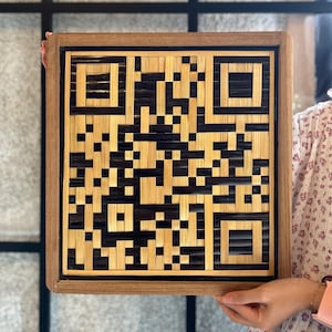 Puede incluir: Una obra de arte cuadrada enmarcada en madera con un código QR negro y marrón hecho de bambú tejido.
