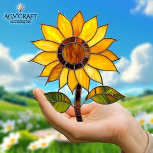Puede incluir: Un girasol de vidrieras con p&eacute;talos amarillos y un centro marr&oacute;n, sostenido en una mano. El tallo y las hojas son verdes y marrones. El logotipo de AGV CRAFT est&aacute; en la esquina superior izquierda, con el texto "Never Ending Craft".