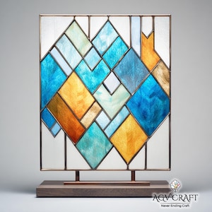 Könnte beinhalten: Eine Glasmalerei mit einem geometrischen Design in Blau-, Türkis- und Goldtönen, in einem quadratischen Rahmen. Das Panel ist auf einem Holzsockel mit Metallstützen montiert. Das Design zeigt Diamanten- und Rechteckformen.