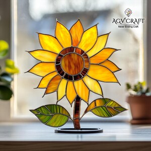 Puede incluir: Un girasol de vidrieras con p&eacute;talos amarillos, un centro marr&oacute;n y hojas verdes. La obra de arte se exhibe sobre un soporte negro. El logotipo "AGV CRAFT Never Ending Craft" es visible en la esquina superior derecha.
