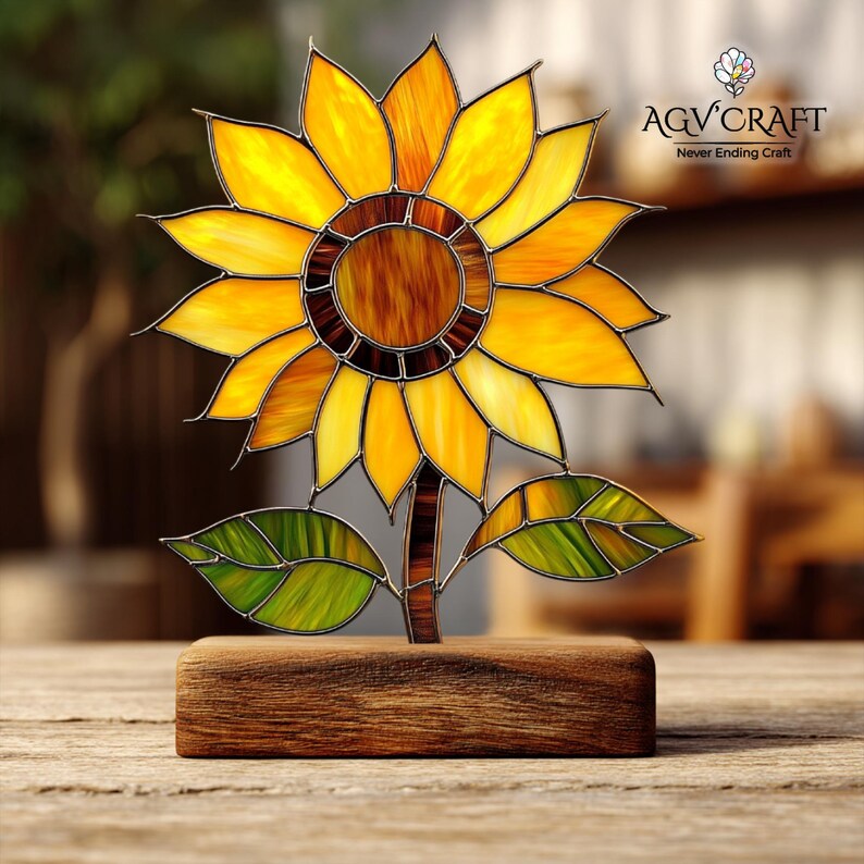 Puede incluir: Una escultura de girasol de vidrieras con p&eacute;talos amarillos, naranjas y marrones, hojas verdes y un tallo marr&oacute;n. La escultura est&aacute; montada sobre una base de madera. El texto "AQV Craft Never Ending Craft" es visible.