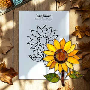 Puede incluir: Un girasol de vidrieras con p&eacute;talos amarillos, naranjas y marrones, hojas verdes y un tallo marr&oacute;n. Un dibujo lineal en blanco y negro de un girasol est&aacute; al lado de la flor de vidrieras. El texto en el papel dice "Sunflower Stained Glass Pattern."