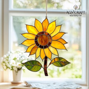 Puede incluir: Un atrapasoles de girasol de vidrieras con p&eacute;talos amarillos, naranjas y marrones, hojas verdes y un tallo marr&oacute;n. El atrapasoles est&aacute; colgado frente a una ventana. El texto "AGV Craft Never Ending Craft" es visible.