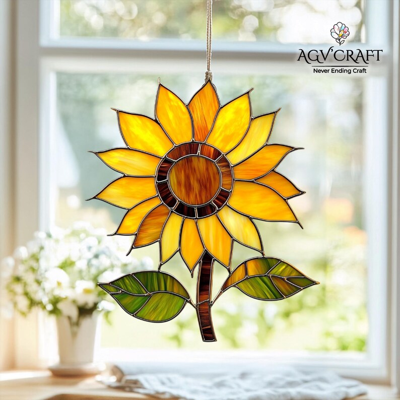 Puede incluir: Un atrapasoles de girasol de vidrieras con p&eacute;talos amarillos, naranjas y marrones, hojas verdes y un tallo marr&oacute;n. El atrapasoles est&aacute; colgado frente a una ventana. El texto "AGV Craft Never Ending Craft" es visible.