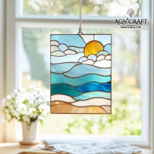 May include: Stained glass panel in a rectangular shape, featuring a seascape with a yellow sun, blue sky, and ocean waves in shades of blue and teal. The panel is framed in a dark metal and suspended by a natural fibre cord. The text "AGV Craft Never Ending Craft" is visible.