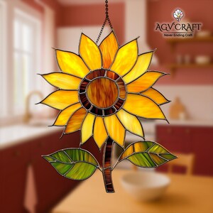 Puede incluir: Girasol de vidriera con p&eacute;talos amarillos, naranjas y marrones, hojas verdes y un tallo marr&oacute;n. El girasol cuelga de una cadena. El logotipo "AGV Craft Never Ending Craft" est&aacute; en la esquina superior derecha.