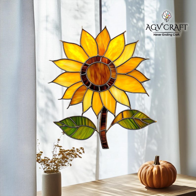 Puede incluir: Un girasol de vidriera en tonos amarillos, naranjas y marrones, con hojas verdes y un tallo marr&oacute;n. El logotipo de AGV Craft es visible. El girasol cuelga frente a una cortina blanca. Objeto decorativo.