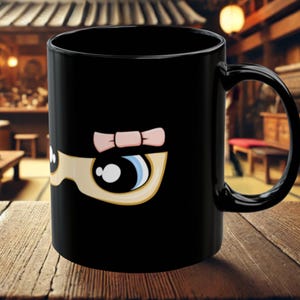 Puede incluir: Taza de cerámica negra con un diseño de ojo de dibujos animados y un lazo rosa. El ojo tiene un iris azul y un contorno marrón claro. La taza está sobre una superficie de madera.