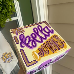 Op de afbeelding: Een vierkante houten doos geschilderd in crème, paars en goud. De bovenkant toont de naam Bella Joyner in paars en goud cursief schrift, met een klein geschilderd vuurwerk. De zijkant toont de tekst Benton High School in zwarte cursieve letters.