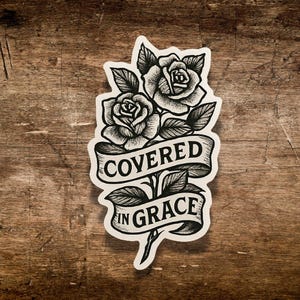 May include: Black and white sticker featuring two roses with leaves and the words "COVERED IN GRACE" on a banner. The sticker has a detailed, illustrative style, with a vintage aesthetic. The background is a wooden surface.