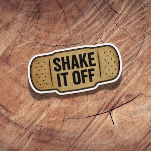May include: A bandage-shaped sticker with the words "SHAKE IT OFF" in bold black letters. The sticker has a tan background with small brown dots, and a white border. The sticker is placed on a brown, textured wooden surface.