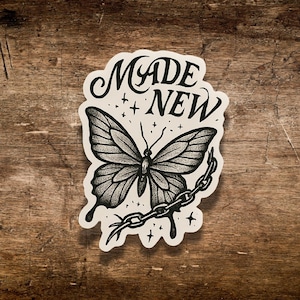 May include: A black and white sticker featuring a detailed butterfly illustration with a chain and the words "MADE NEW". The sticker has a white border and is set against a wooden background.