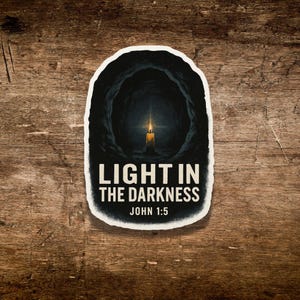 May include: A sticker with a black and white design featuring a candle in a dark cave. The text "LIGHT IN THE DARKNESS" and "JOHN 1:5" are displayed in white. The sticker is set against a wooden background.