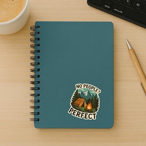 May include: A blue notebook with a spiral binding and a sticker that says "No people? Perfect." The sticker features a cartoon illustration of a campsite with a tent, a campfire, and mountains in the background.