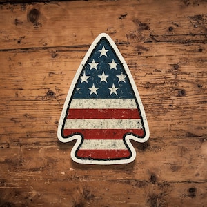 May include: A red, white, and blue arrowhead-shaped sticker with a distressed American flag design. The sticker is on a wooden background.