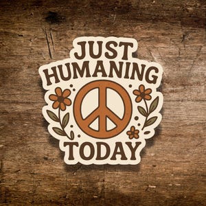May include: A sticker with the text "JUST HUMANING TODAY" in a retro font. The design features a peace sign in the center, surrounded by flowers and leaves in shades of brown, orange, and green. The sticker is on a wooden surface.