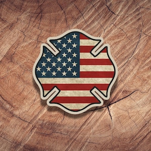 May include: A distressed-style firefighter badge featuring the American flag. The badge is in the shape of a Maltese cross, with the stars and stripes of the US flag filling the center. The badge has a white border and is set against a wood grain background.