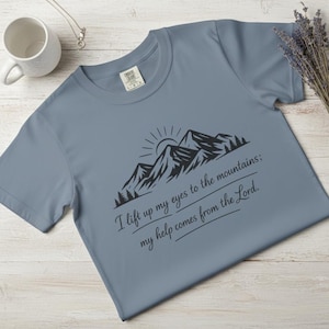 May include: A dusty blue t-shirt featuring a black mountain graphic with a rising sun and the text "I lift up my eyes to the mountains; my help comes from the Lord." The shirt has a crew neck and short sleeves.