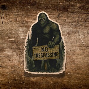 May include: A sticker featuring a Bigfoot illustration holding a "NO TRESPASSING" sign. The Bigfoot is depicted in shades of green and black, with glowing yellow eyes. The background includes stylized trees, set against a rustic wood grain.