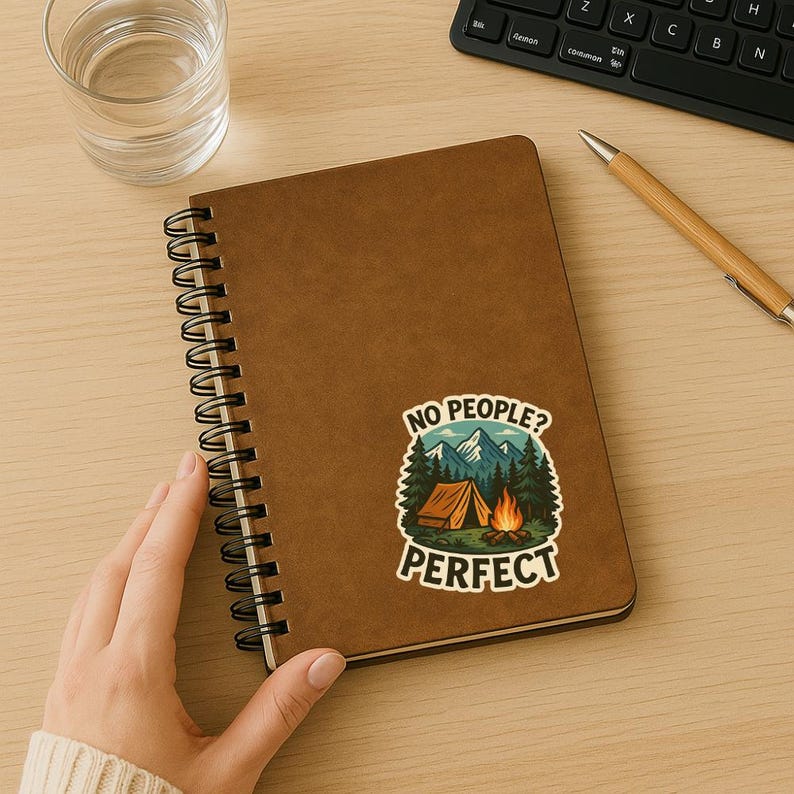 May include: A brown spiral notebook with a sticker on the front. The sticker features a cartoon illustration of a campsite with a tent, a campfire, and mountains. The text on the sticker reads "No People? Perfect."