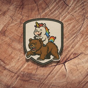 May include: Embroidered patch featuring a cartoon unicorn riding a brown bear. The unicorn has a rainbow mane and tail, and the bear has an angry expression. The patch has a white background with a black border.