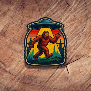 May include: A colorful sticker featuring a Bigfoot running away from a UFO. The design includes a sunset background with trees and a bright light shining down on the creature. The sticker has a dark blue border.