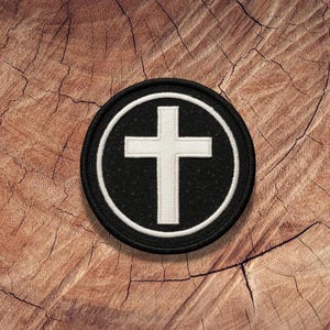May include: A black and white embroidered patch with a white cross in the center. The cross is surrounded by a black circle with a white outline.