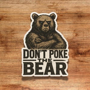 May include: A sticker featuring a stern-faced bear with crossed arms. The text "DON'T POKE THE BEAR" is printed below the bear in a bold, distressed font. The sticker has a vintage, rustic aesthetic.