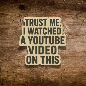 May include: A rectangular sticker with the text "TRUST ME, I WATCHED A YOUTUBE VIDEO ON THIS" in a bold, black font. The sticker has a light brown background and is placed on a wooden surface.