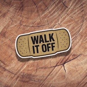 May include: A tan bandage sticker with the words "WALK IT OFF" in bold black letters. The sticker has a white border and small dotted details. The background is a close-up of a brown, textured wood surface.