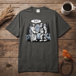 May include: Dark grey t-shirt with a graphic of Jesus and Bigfoot shaking hands, with the text "I WAS WAITING FOR YOU..." in a speech bubble. The design is set against a backdrop of trees and mountains.